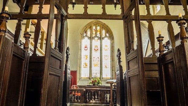 Blick ins Innere einer Kirche mit Holzbänken, einem Glasfenster und einer Blumenkomposition auf einem Podest.