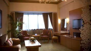 Habitación de hotel con zona de comedor, dos sillones, sofá, televisión y un gran ventanal con cortinas.