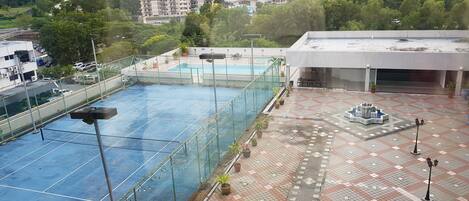 Una cancha de tenis en la azotea con red, rodeada de plantas en macetas y un área de descanso con mesas y sillas.