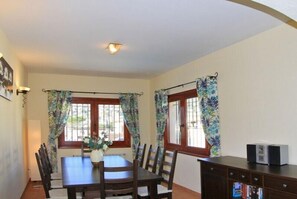 A dining area with a table and chairs, two windows with curtains, a wall-mounted light fixture, and a small cabinet with a television. - Club Villamar - Nice villa with airco, wifi, fenced private swimming pool nearby Calonge (Calonge)