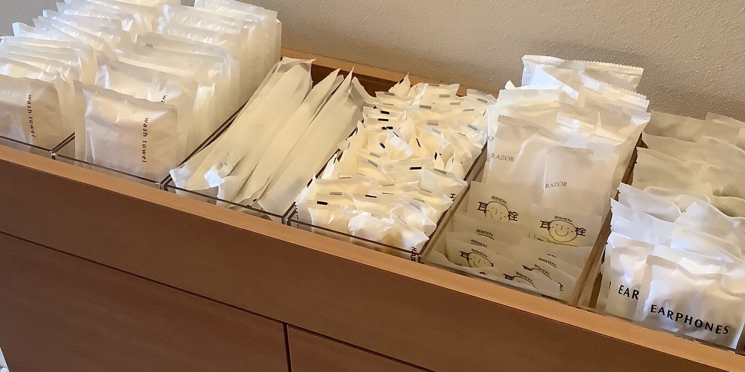 Un espacio de almacenamiento con varios compartimentos que contienen bolsas blancas etiquetadas con artículos como 'toalla de baño', 'afeitadora', 'auriculares' y 'tapones para los oídos'.