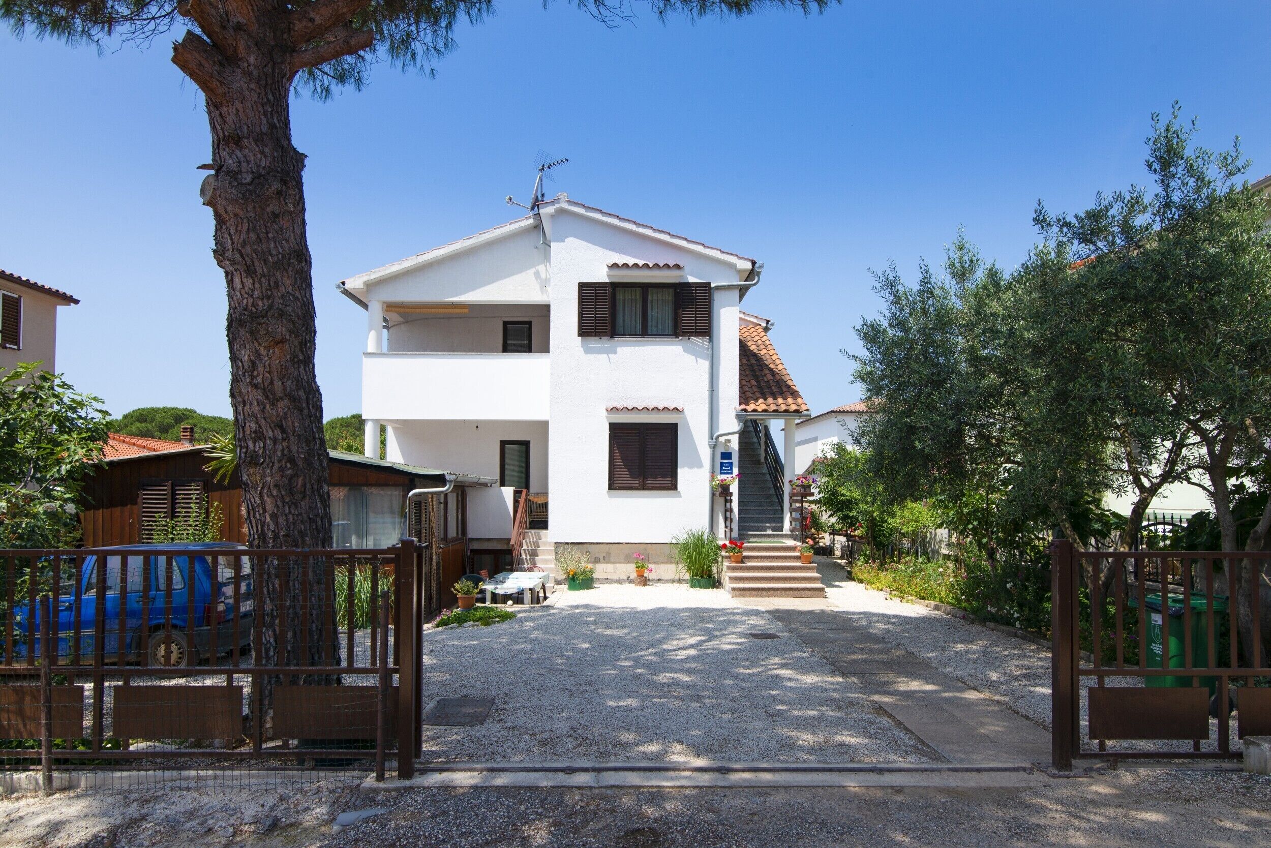 A two-story white building with a tiled roof, a staircase leading to the entrance, and a gravel driveway.
