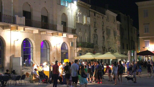 A crowded square with historic buildings, outdoor seating, and umbrellas.