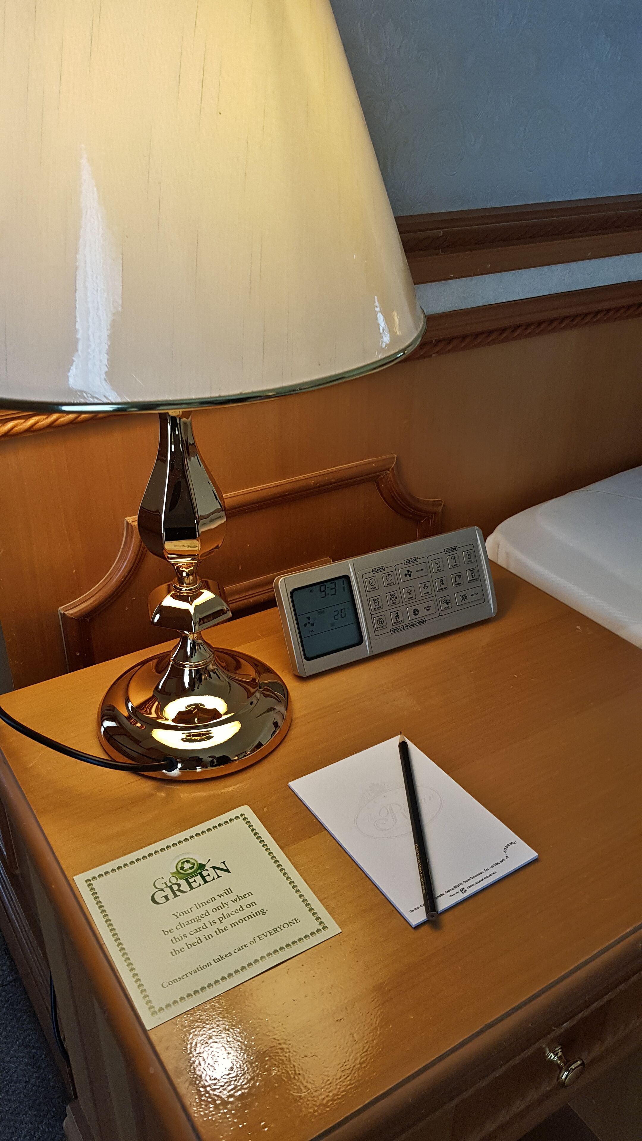 A wooden nightstand with a white lamp, a digital clock, a notepad, and a pen.