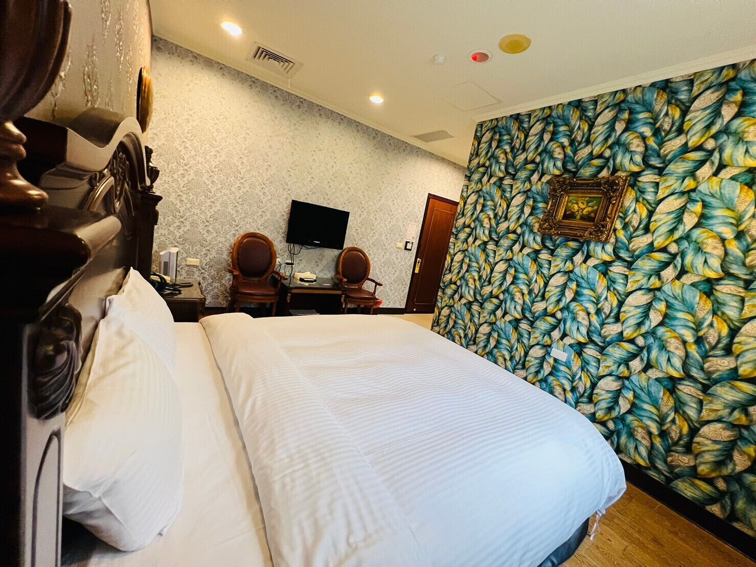 Habitación de hotel con una cama grande, dos sillas de madera, un escritorio con televisor y una pared con diseño.