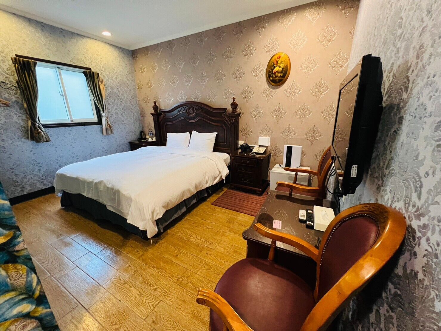 Hotelkamer met een groot bed, een houten nachtkastje, een bureau met stoel, een televisie en behang met een patroon.