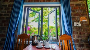 A table set with tea cups, a teapot, and a bottle, with a view of greenery through a window with blue curtains. - Phong Nha An An Homestay (Phong Nha)