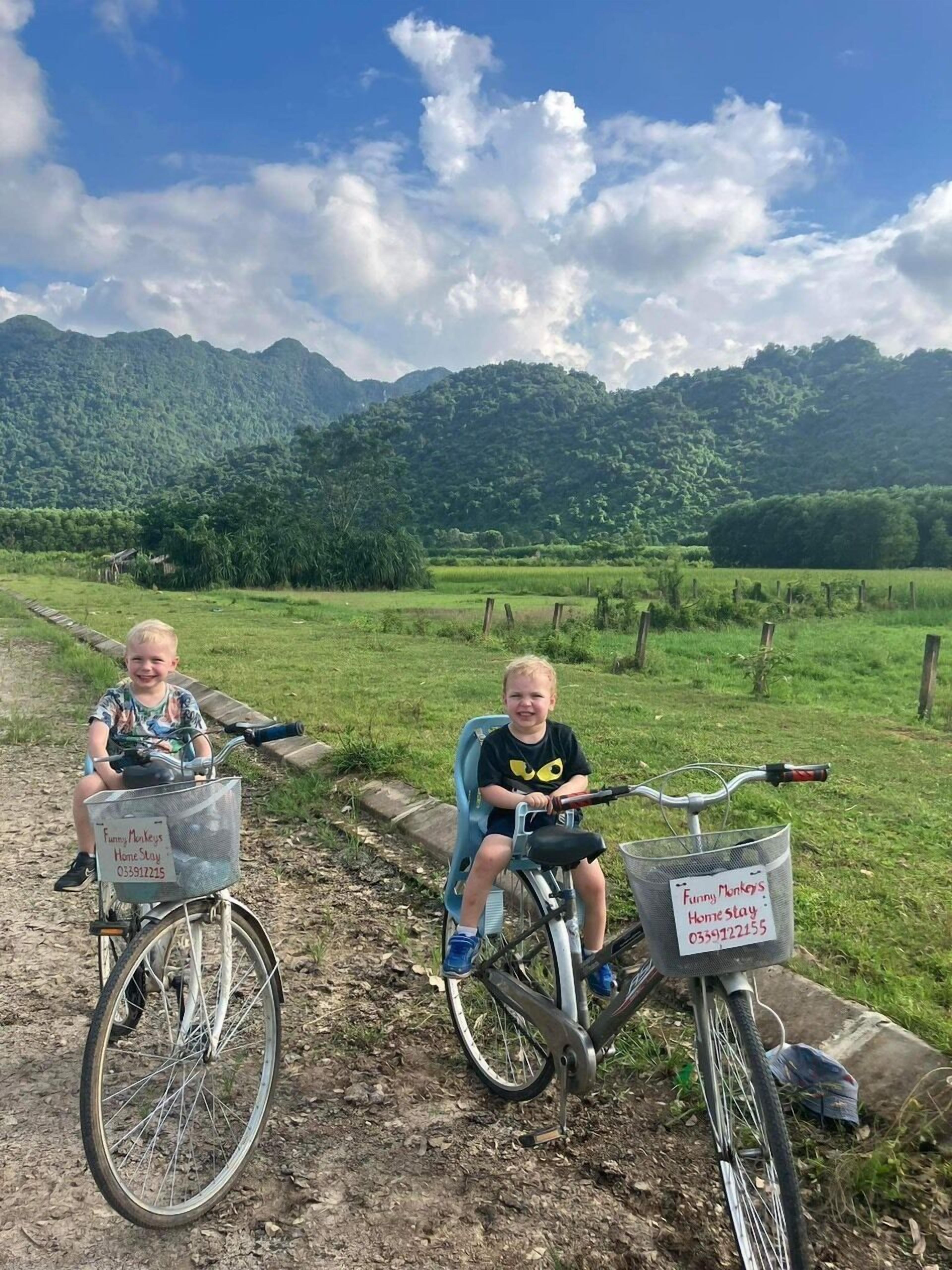 Two children on bicycles with baskets labeled "Funny Monkeys Home Stay" in a rural setting with mountains in the background.