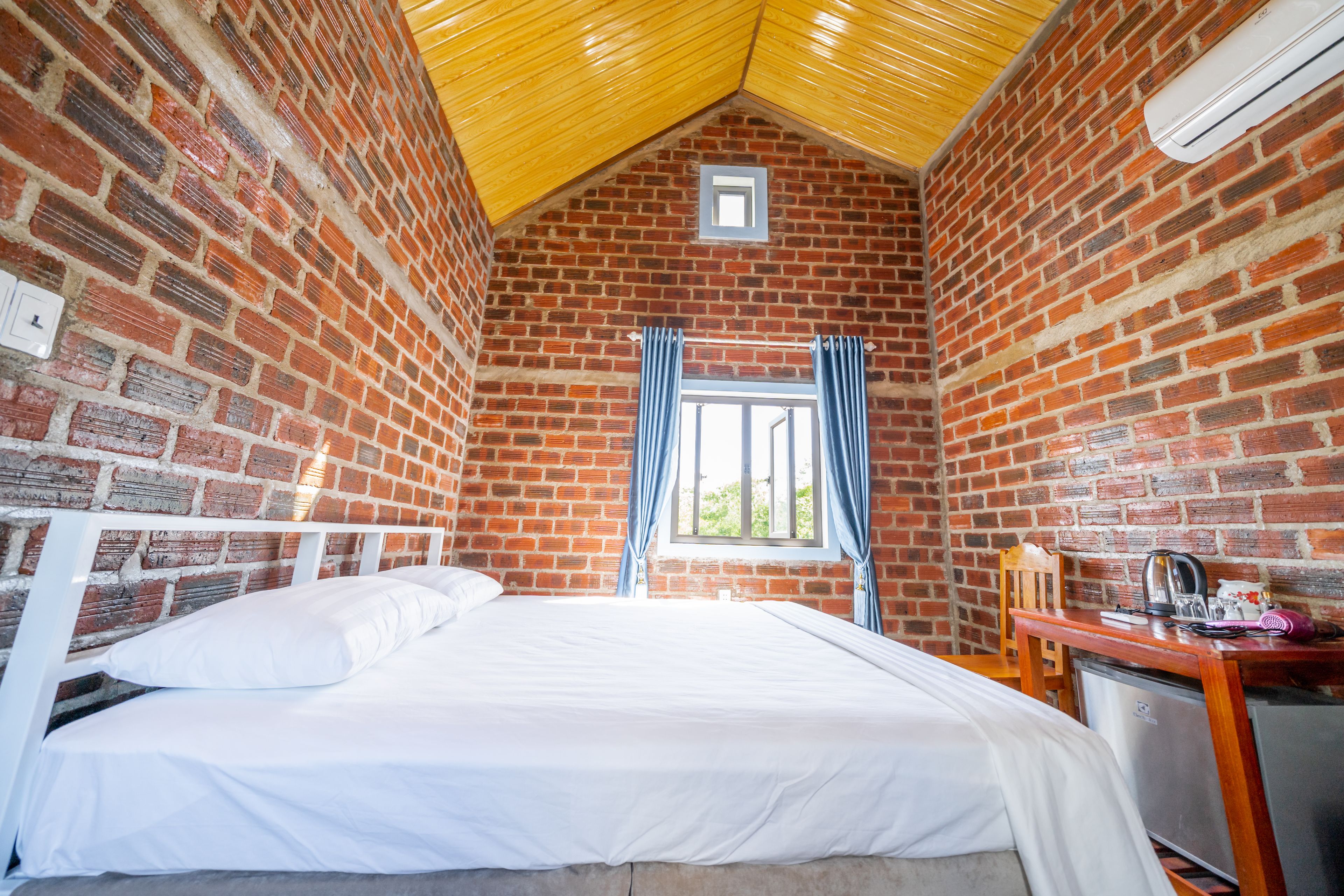 A room with a brick wall, a bed with white bedding, a window with blue curtains, a wooden desk with a kettle, and a small air conditioning unit on the wall.