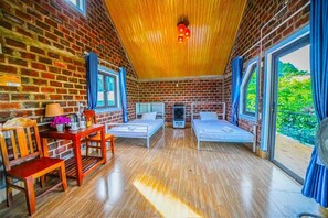 A cozy room with brick walls, wooden ceiling, and large windows. There are two beds, a table with chairs, and a small heater.