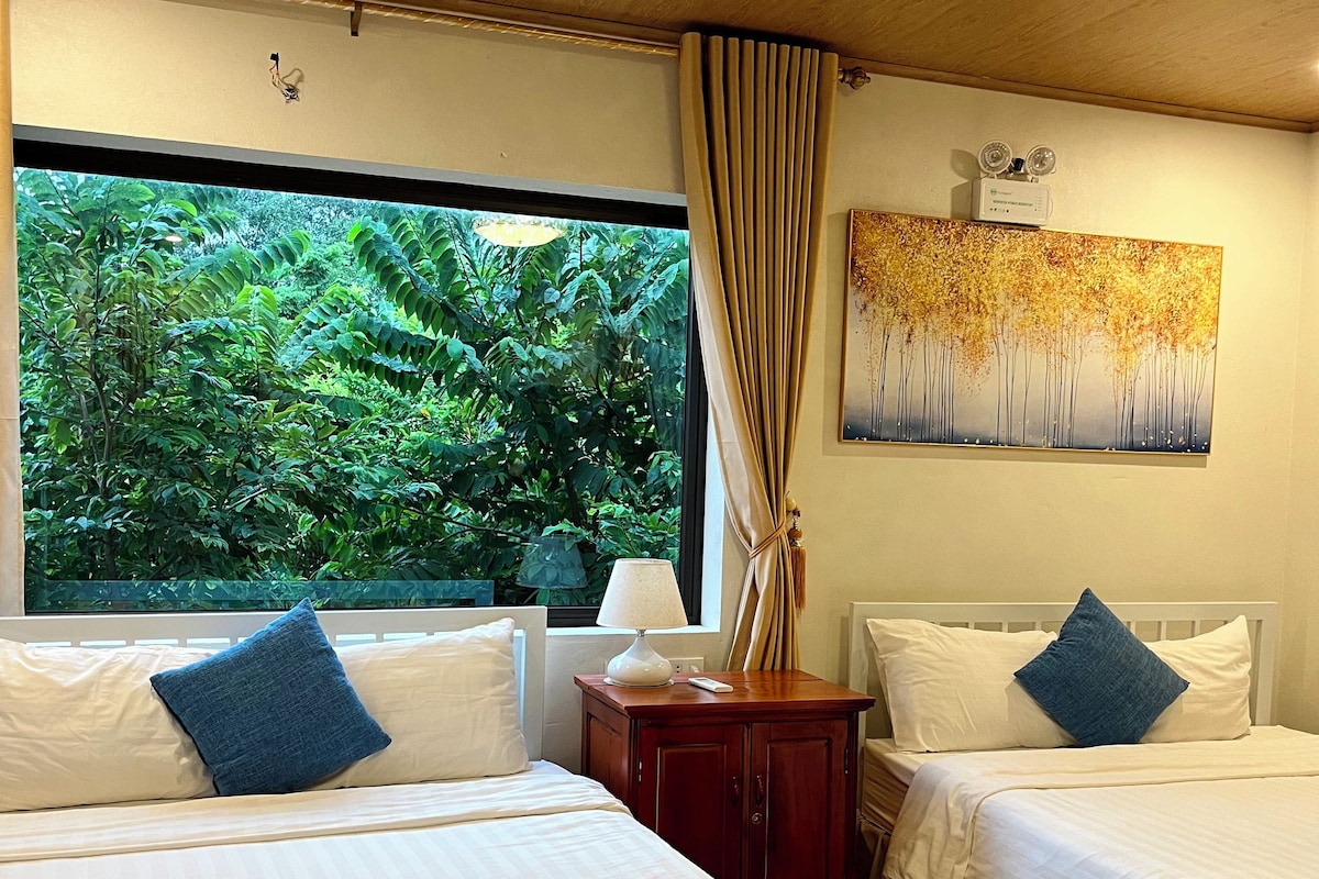 A hotel room with two beds, a large window with a view of greenery, a painting on the wall, and a bedside table with a lamp.