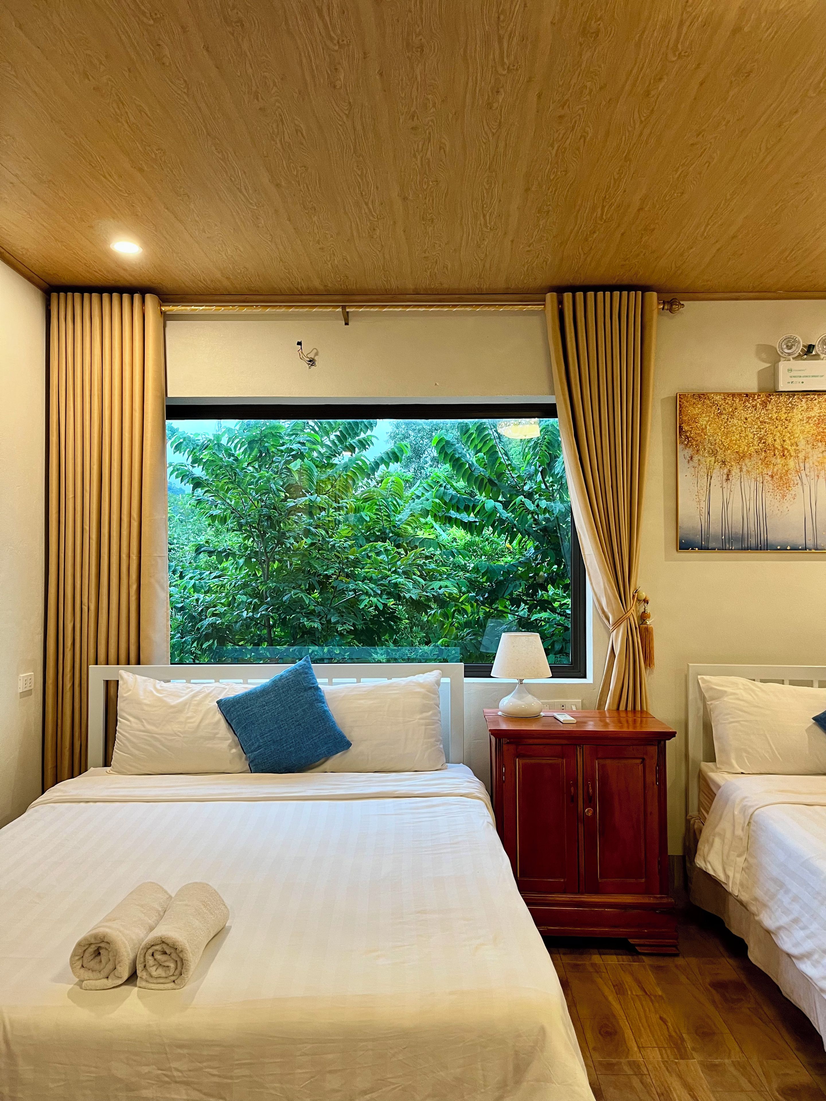 A hotel room with a large window, a bed with white linens, a bedside table with a lamp, and a wooden headboard.