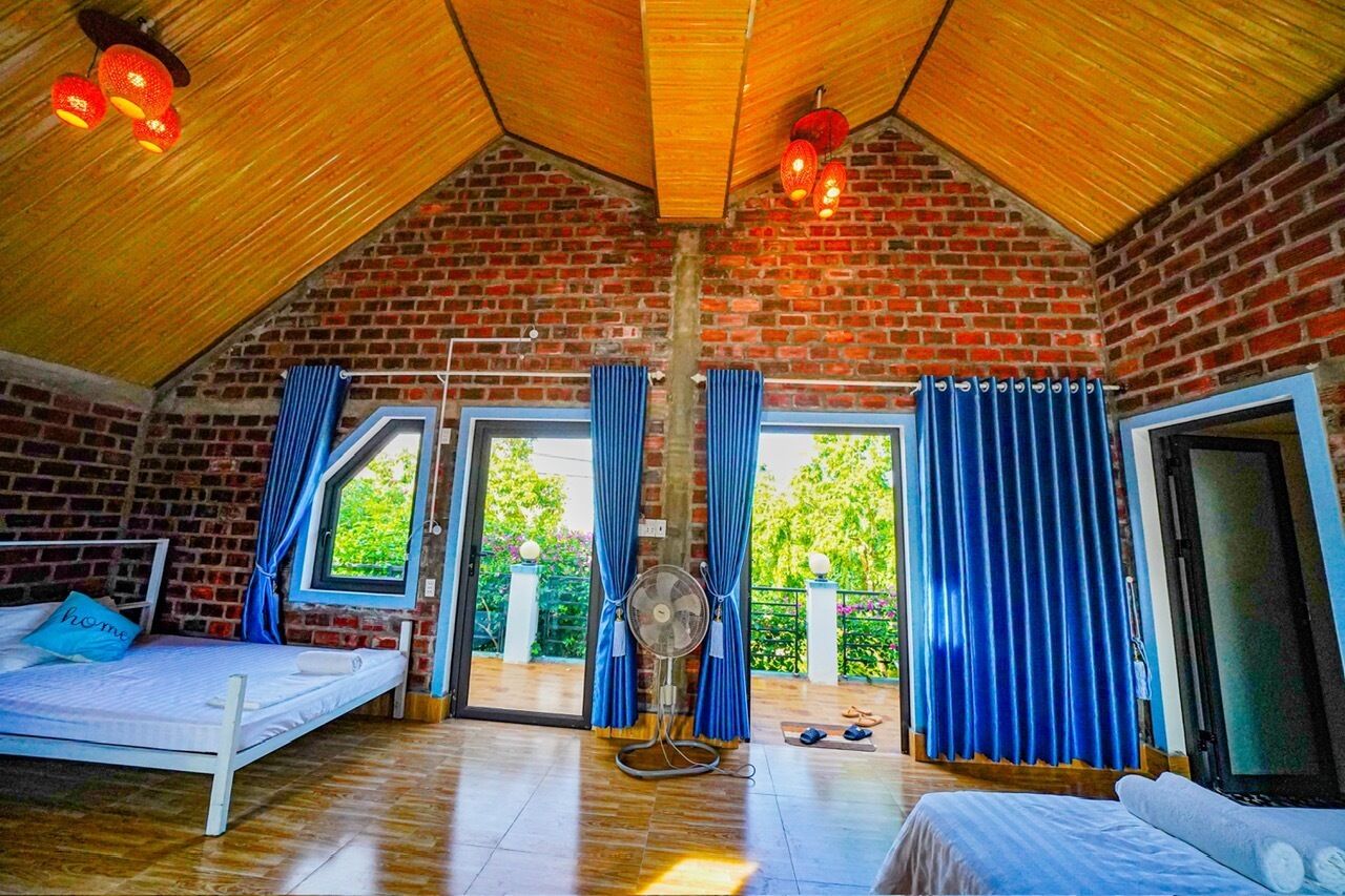 Una habitación con techo inclinado, paredes de ladrillo y un ventanal con cortinas azules. Hay dos camas con estructura blanca y almohadas azules.