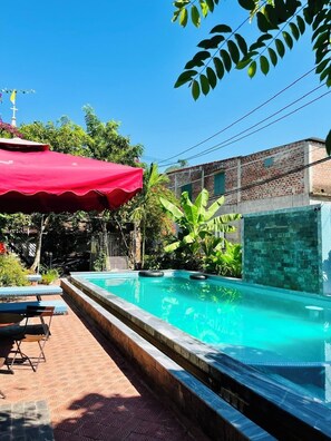 A swimming pool with a red umbrella, surrounded by greenery and a brick building.