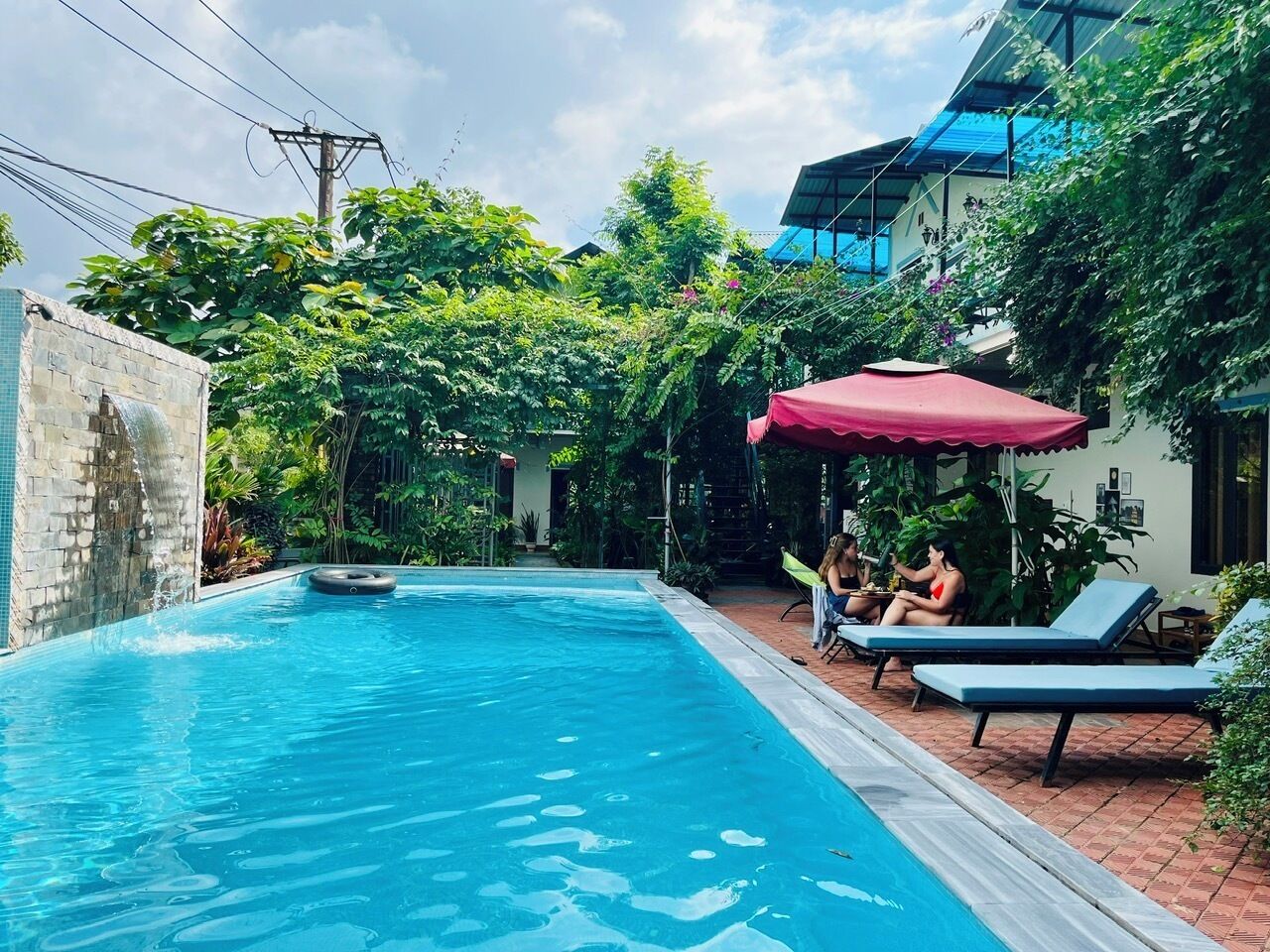 Una piscina con sillones de descanso y una sombrilla roja, rodeada de vegetación exuberante y un edificio con techo azul.