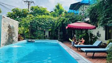 Una piscina con sillones de descanso y una sombrilla roja, rodeada de vegetación exuberante y un edificio con techo azul.