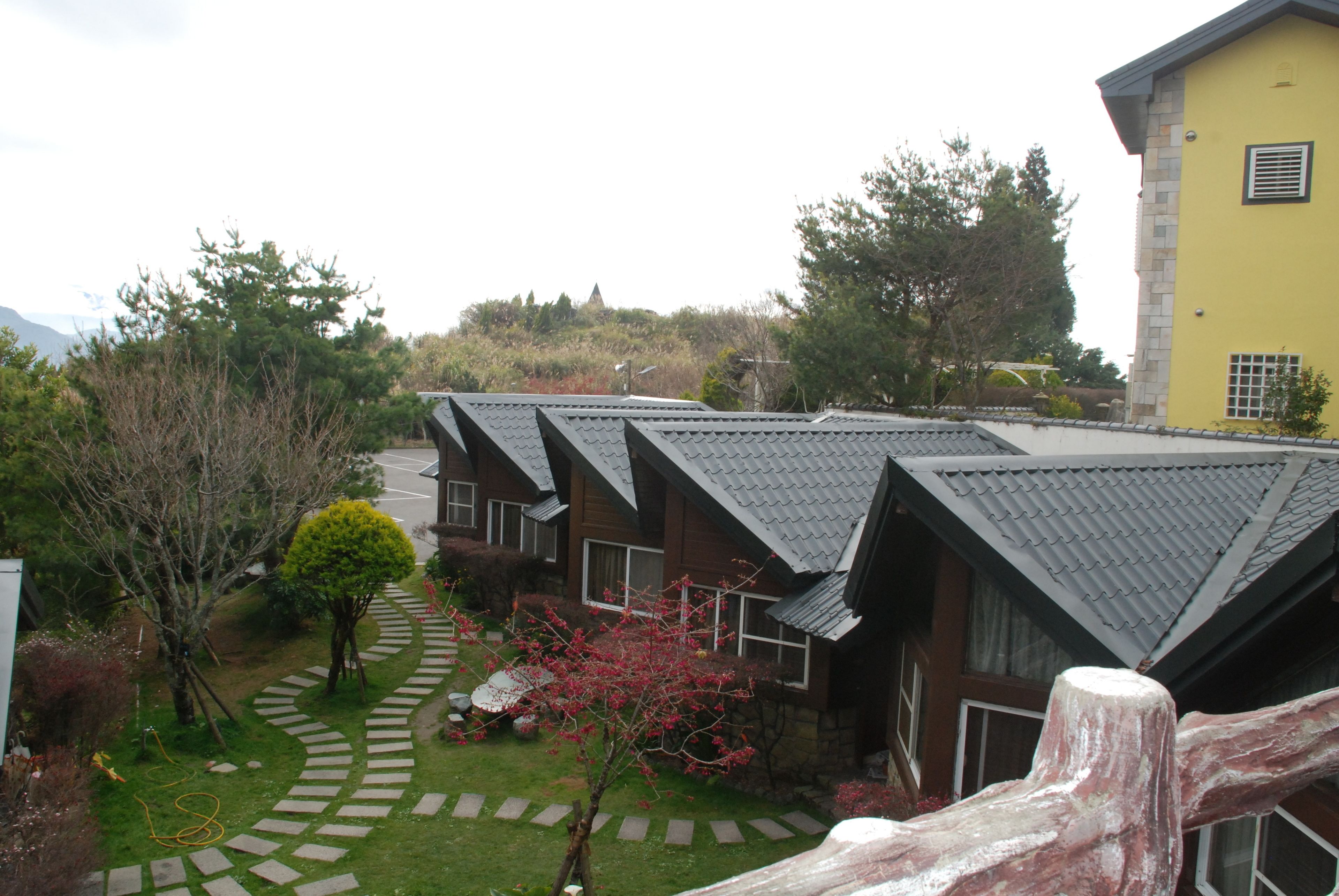 A row of modern houses with sloped roofs, a paved pathway, and a small garden area with a tree and shrubbery.