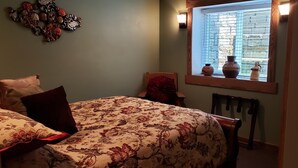 Ein Schlafzimmer mit floralem Bettüberwurf, einem Holzstuhl, einem Fenster mit Dekoration und einer Wandleuchte.