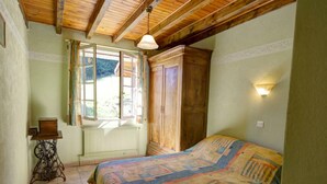 A bedroom with a wooden ceiling, a bed with a colorful quilt, a radiator, a sewing machine, and a window with curtains. - Authentic cottage with fireplace, pets allowed and WiFi by the forest (La Versanne)
