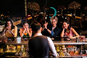 Eine Bar mit einem Barkeeper, der Getränke zubereitet, einer Theke mit verschiedenen Flaschen und Gläsern, und einem Stadtblick im Hintergrund.