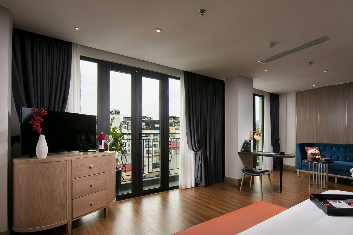 A modern hotel room with a large sliding glass door to a balcony, a flat-screen TV, a wooden dresser, a blue sofa, and a small dining table.