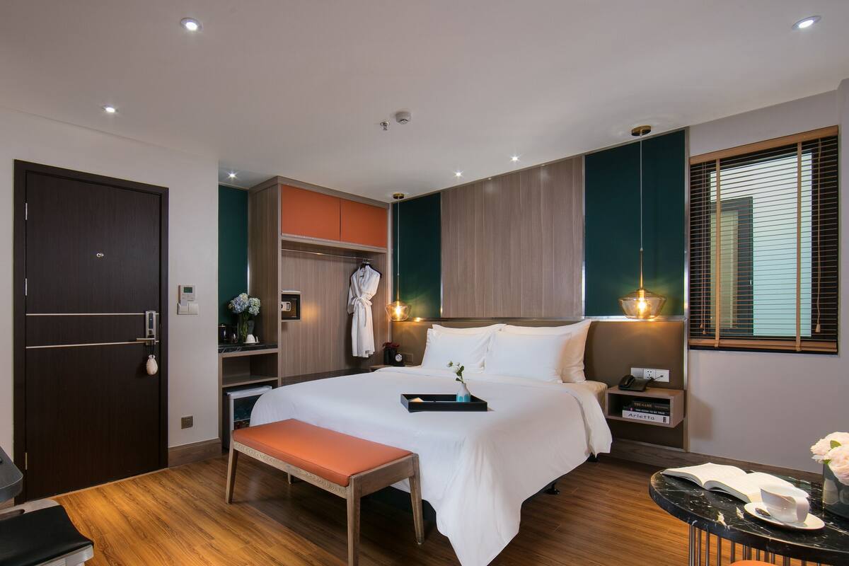 A modern hotel room with a large bed, bedside table, desk, and a window with blinds.