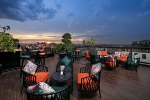 A rooftop terrace with wicker chairs and tables, a view of the city skyline, and a sunset in the background. - Aurora Premium - A Lifestyle Hotel (Hanoi)