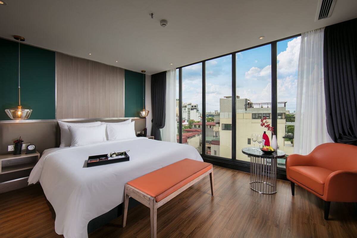 A modern hotel room with a large bed, a bench, an orange armchair, a small round table, and a view of the city through large windows.