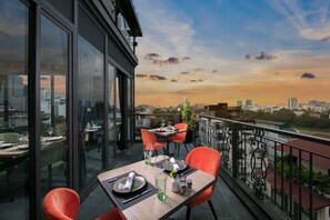 A dining area with a table set for two, overlooking a cityscape at sunset. - Aurora Premium - A Lifestyle Hotel (Hanoi)