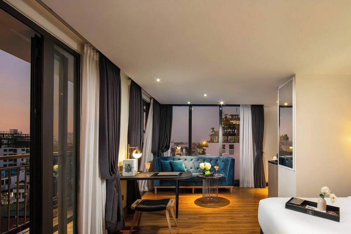 A hotel room with a large window, a bed, a seating area with a blue sofa, and a dining table.