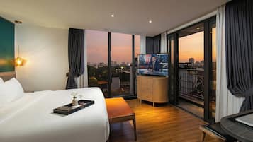 A hotel room with a bed, a desk with a laptop, a TV, and a balcony with a city view.