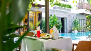 A dining table set with plates, a glass of red beverage, and a bottle of orange juice, overlooking a swimming pool. - December Hoi An Villa (Da Nang)