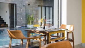 A dining area with a wooden table and chairs, a vase with flowers, and a view of a swimming pool and outdoor seating area. - December Hoi An Villa (Da Nang)