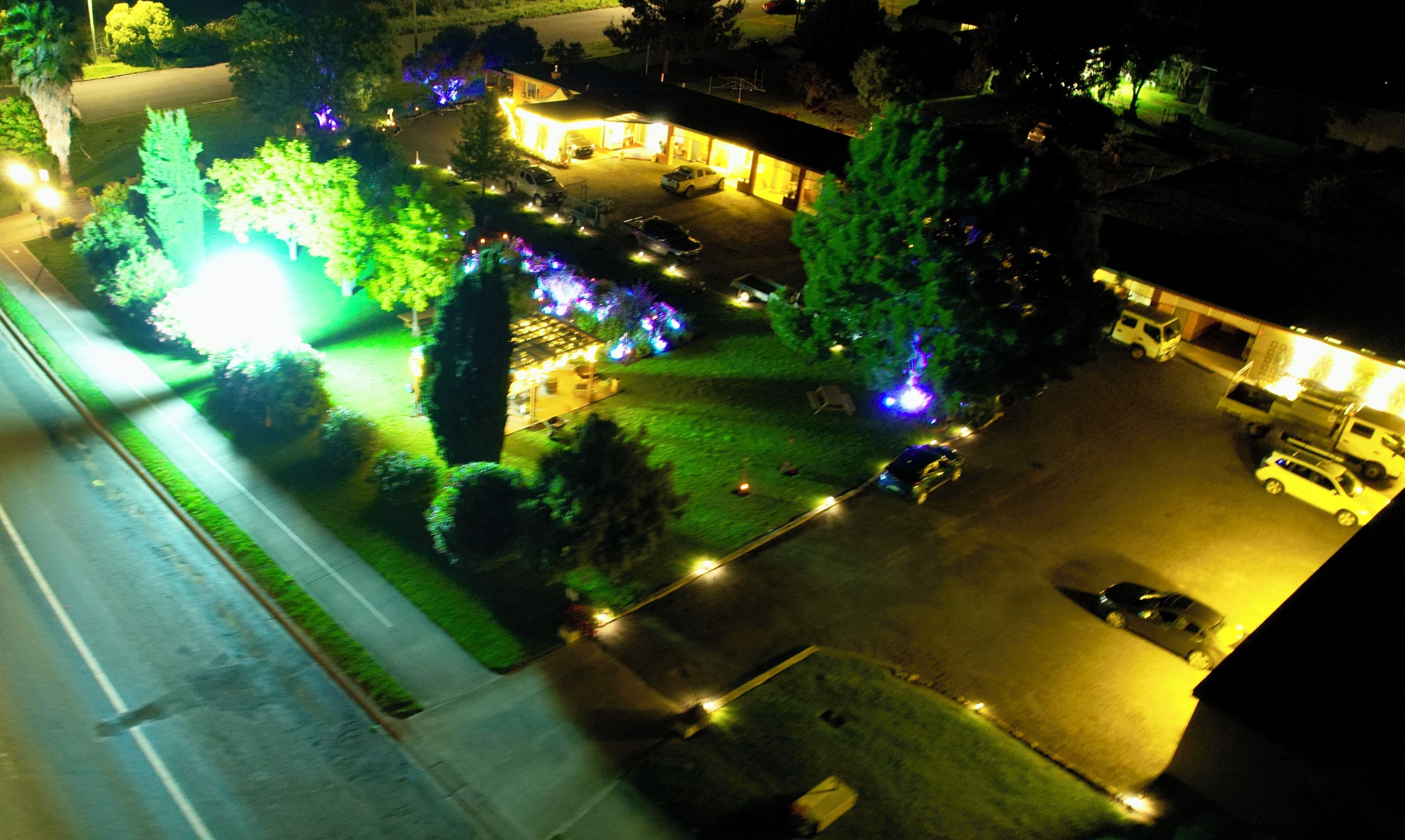 Vista aérea de um estacionamento com vários carros, um edifício com janelas iluminadas e vegetação ao redor.
