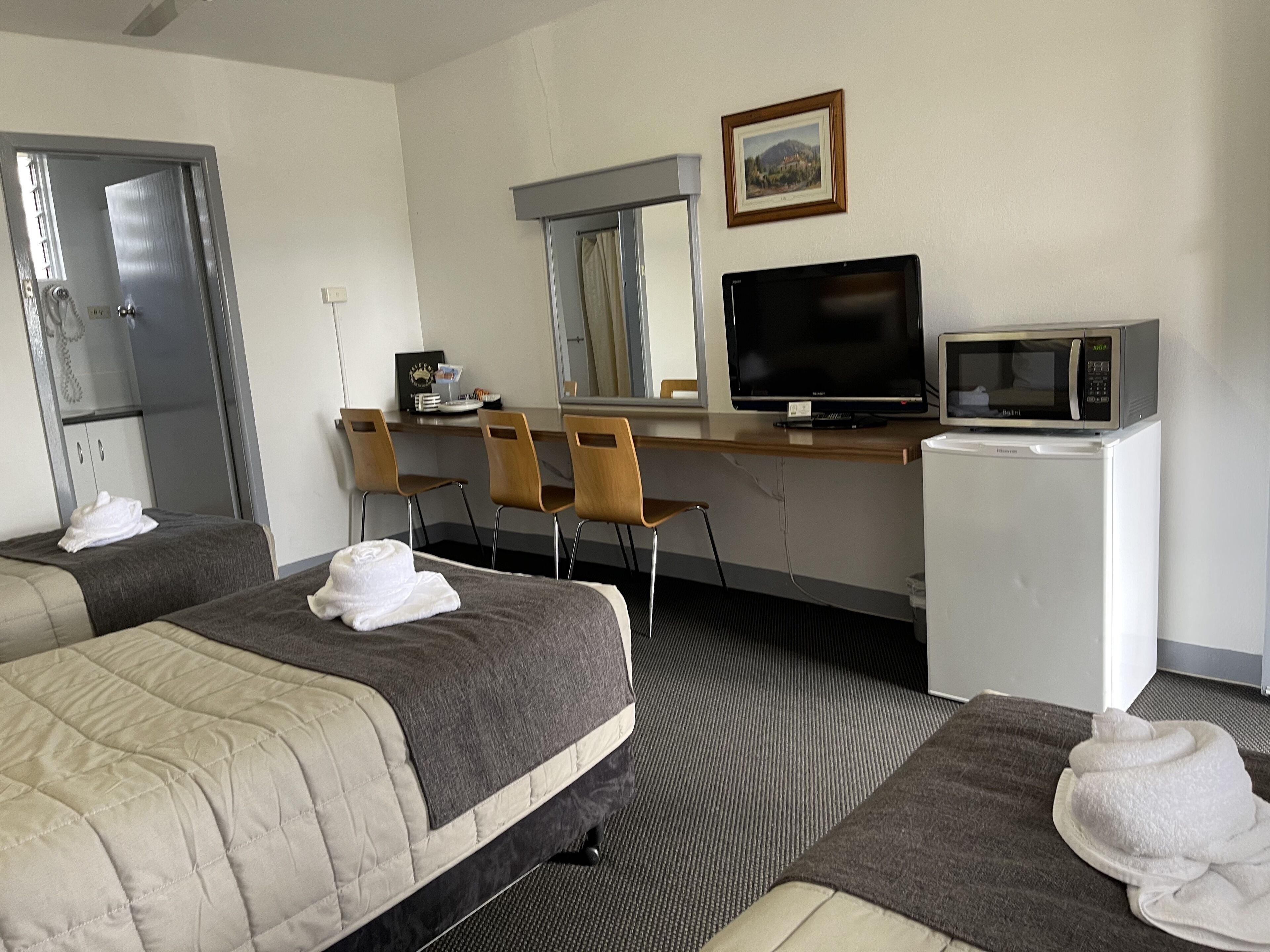 Ein Hotelzimmer mit zwei Betten, einem Schreibtisch mit Fernseher und Mikrowelle, einem kleinen Kühlschrank und einem Fenster mit Blick auf eine Landschaft.