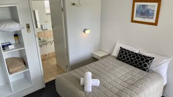 Une chambre d’hôtel comprenant un lit, une table de chevet, une commode, une étagère avec des articles de toilette, une serviette sur le lit, un tableau encadré au mur et un appareil de climatisation.