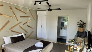 Quarto com cama, escrivaninha e cadeira. Armário e banheiro visíveis por uma porta aberta.
