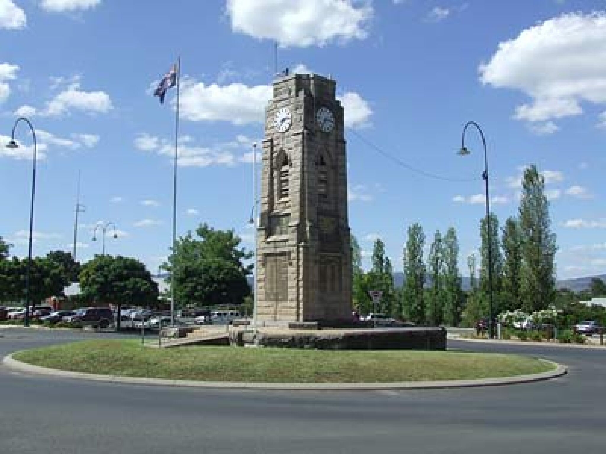 A stone clock tower with a clock face on each side, surrounded by a grassy area and street lamps.