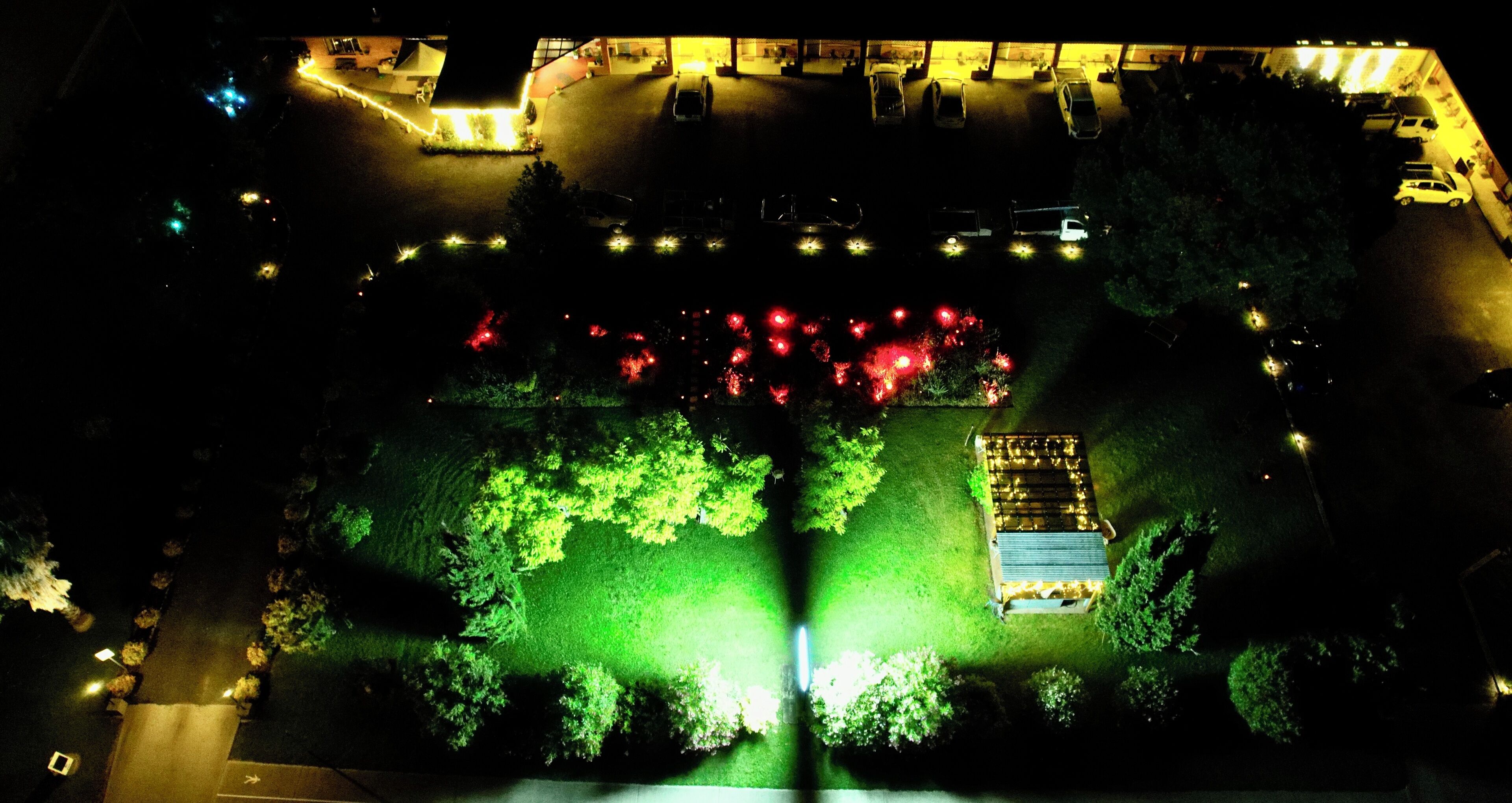 Vista aérea de un estacionamiento con varios autos, una zona iluminada con árboles y un edificio de techo inclinado.