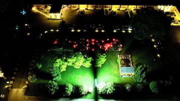 Vista aérea de un estacionamiento con varios autos, una zona iluminada con árboles y un edificio de techo inclinado.