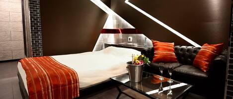 A modern hotel room with a bed, a glass coffee table, a sofa, and a fireplace.