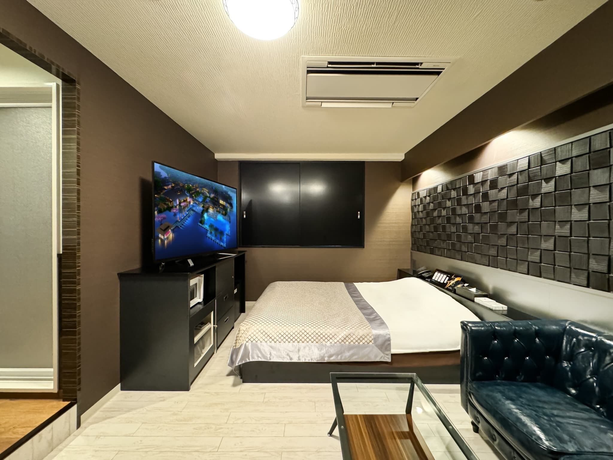 A modern hotel room with a large bed, a TV mounted on the wall, a dark-colored headboard, and a leather sofa.