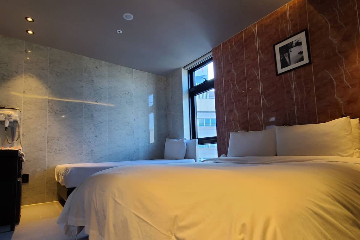 Habitación de hotel con una cama grande, una mesita de noche pequeña, un secador de pelo y una ventana con vistas a la ciudad.