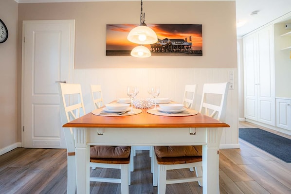 Une salle à manger avec une table en bois dressée pour quatre personnes, des chaises blanches et un luminaire suspendu.