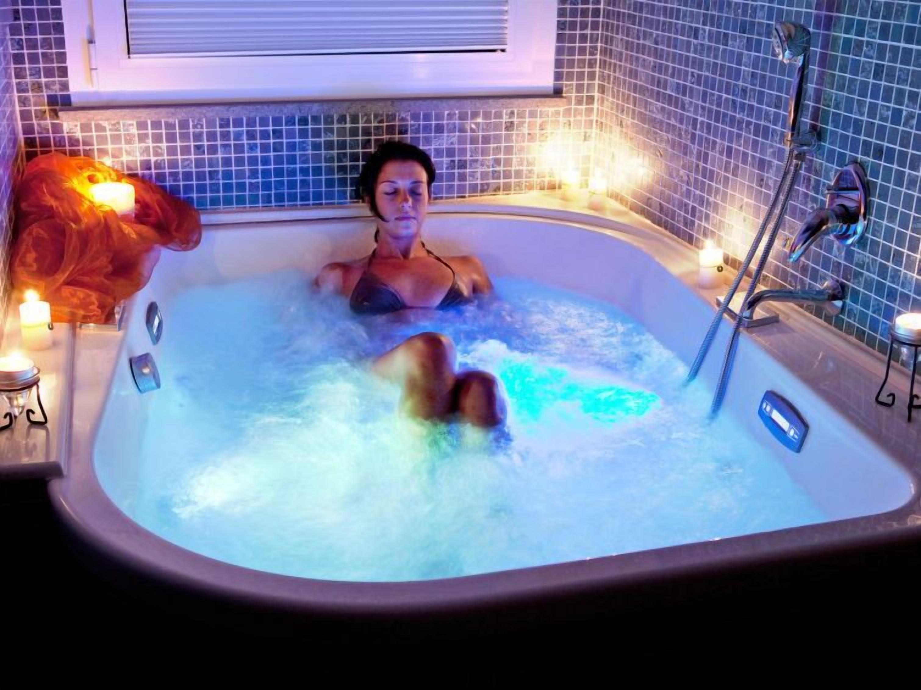 A person relaxing in a hot tub with a control panel, surrounded by tiled walls and candles.