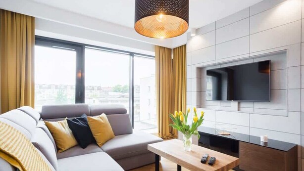 Una sala de estar moderna con un sofá gris, una mesa de centro de madera, un televisor de pantalla plana montado en la pared y un ventanal con vistas a la ciudad.