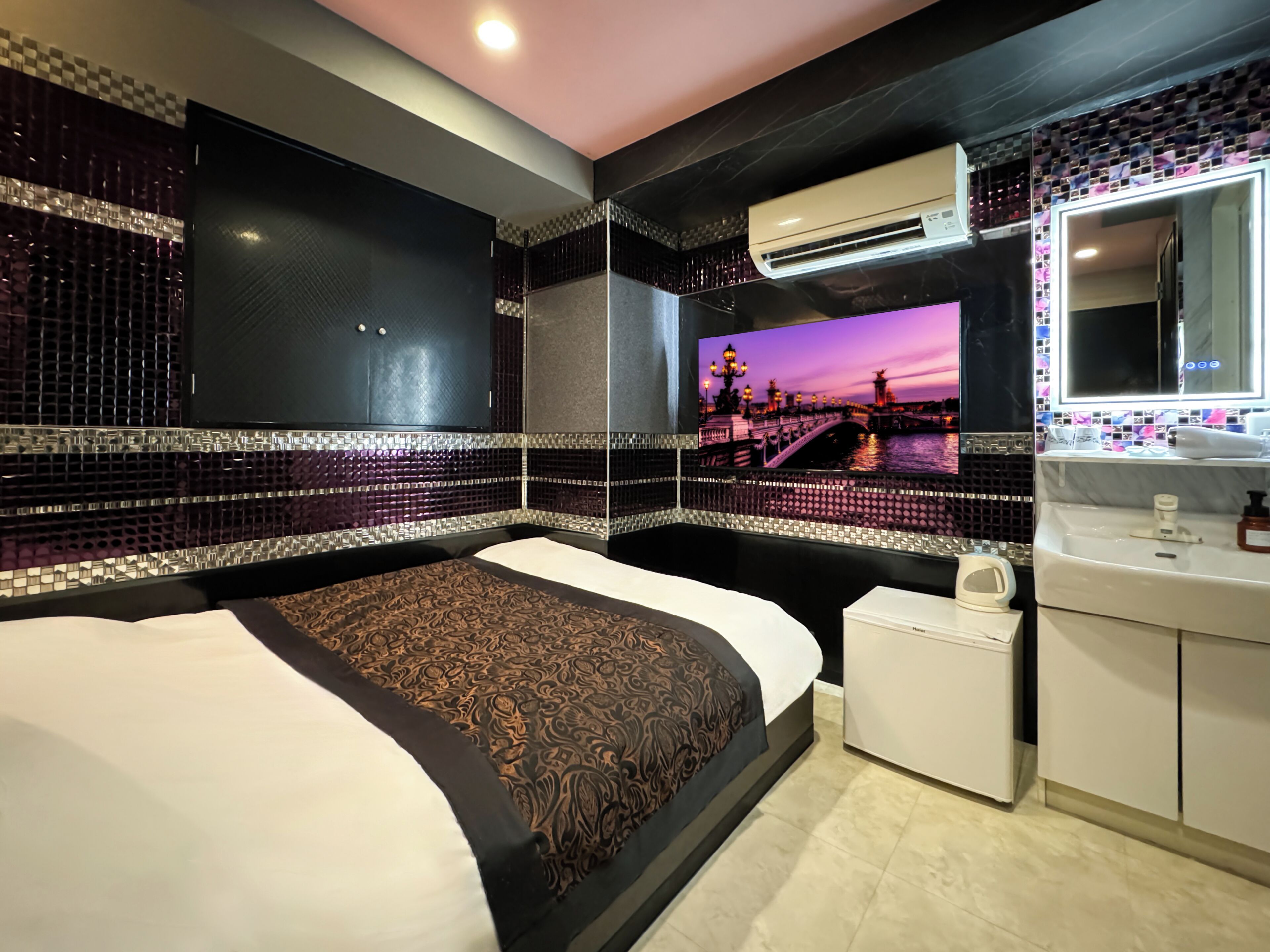 A modern hotel room with a large bed, a small bedside table, a built-in air conditioning unit, a mirror, and a wall-mounted TV displaying a scenic view.
