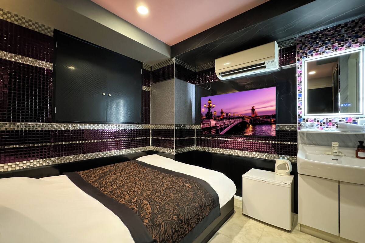 A modern hotel room with a large bed, a small bedside table, a built-in air conditioning unit, a mirror, and a wall-mounted TV displaying a scenic view.