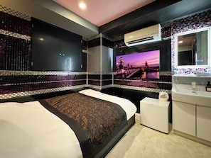 Habitación de hotel moderna con una cama grande, una mesita de noche pequeña, aire acondicionado integrado, un espejo y un televisor de pared que muestra una vista panorámica.