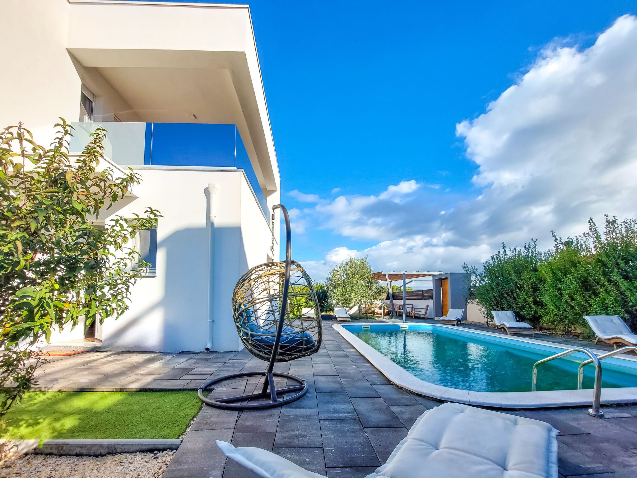Un área de piscina moderna al aire libre con una silla colgante, sillones de descanso y un pequeño edificio.